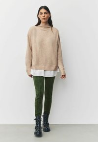 Pull à col roulé beige oversize, chemise blanche à multiples couches en dessous, associé à un pantalon slim vert et des bottes de combat noires à lacets.