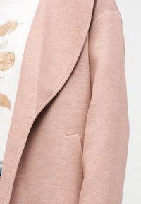 Άτομο που φοράει ανοιχτό ροζ υφασμάτινο σακάκι πάνω από ένα λευκό πουκάμισο με μπεζ εκτυπώσεις φτερών και εμφανές μερικό κείμενο.