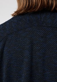 Pull en tricot bleu marine et noir avec un fil métallique subtil. Présente un tissu texturé et un design avec une couture d'épaule arrondie.