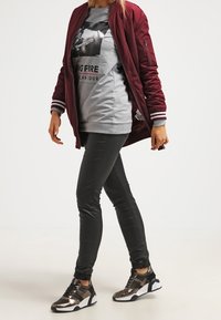 Bordowa kurtka bomber z prążkowanymi mankietami, szary sweter z grafiką, czarne dopasowane dżinsy i sneakersy z metalicznymi akcentami. Swobodny outfit.