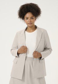 Lichtgrijze pinstripe blazer met een relaxte pasvorm, notch revers en opgestikte zakken. Gecombineerd met een lichtgekleurde top en een rok met elastische taille.