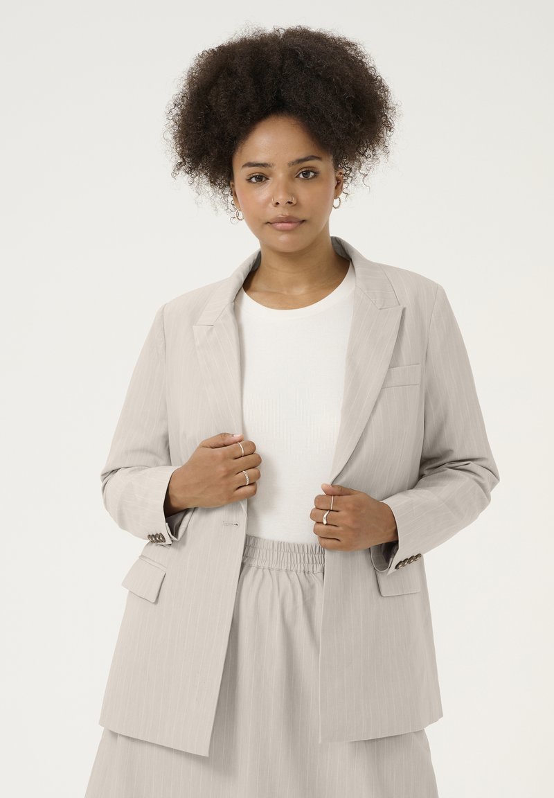 Lichtgrijze pinstripe blazer met een relaxte pasvorm, notch revers en opgestikte zakken. Gecombineerd met een lichtgekleurde top en een rok met elastische taille.