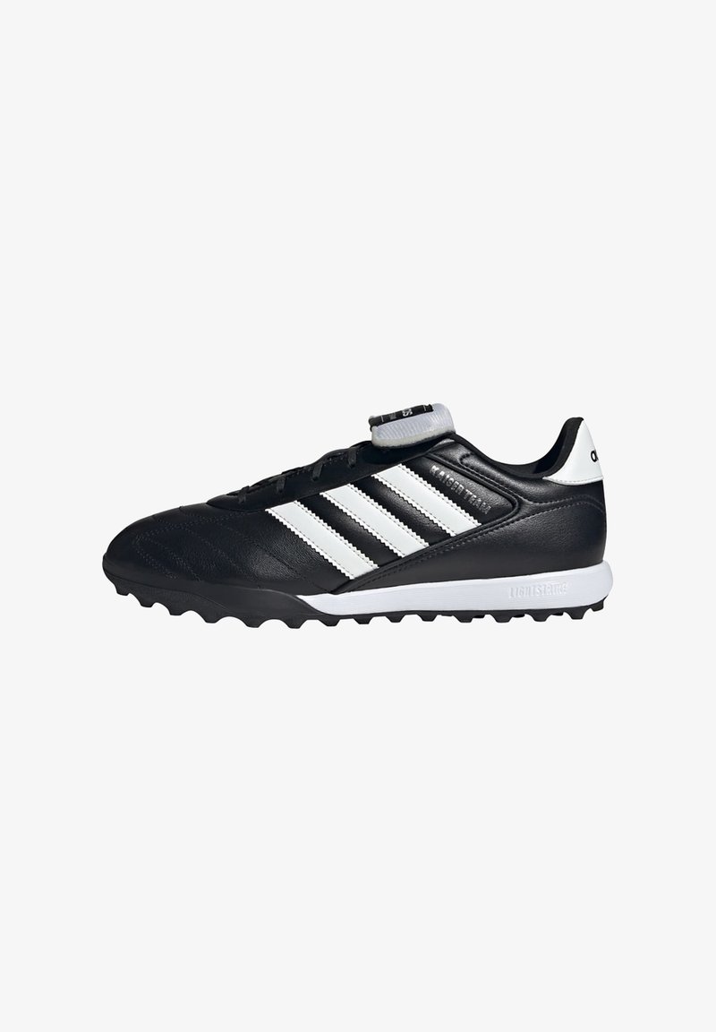 Scarpetta da calcio nera Adidas con strisce bianche, suola strutturata e linguetta pieghevole con logo su sfondo bianco.