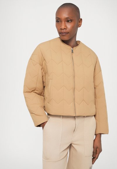 Giacca trapuntata beige con collo rotondo, chiusura a zip e motivo a zig-zag. Il modello indossa pantaloni color crema, che mostrano una vestibilità comoda.