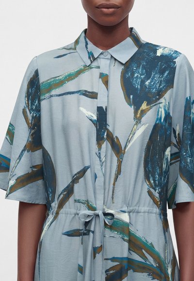 Personne portant une robe bleu clair avec un motif floral abstrait marron, bleu foncé et blanc, col boutonné et détail noué à la taille.