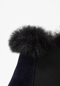 Granatowy zamszowy botek z czarnym futrzanym pomponem u góry oraz kontrastowym, teksturowanym panelem materiałowym po boku.