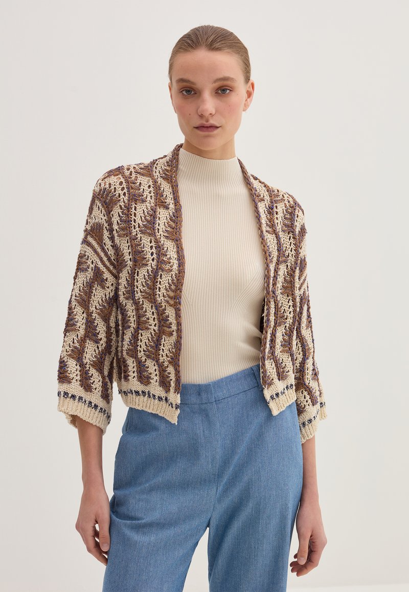 Cardigan lavorato a maglia con un motivo a onde in crema, marrone e blu navy. Maniche a tre quarti e design frontale aperto, abbinato a una maglietta beige e pantaloni blu.