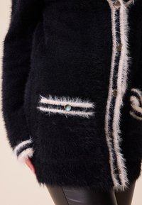 Černý huňatý kardigan s jemným pruhovaným lemováním, se stříbrnými knoflíky a dvěma předními kapsami s dekorativními akcenty.