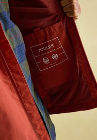 Rokas turēšana atvērta sarkana lietus jaka, kurā redzama iekšējā kabata ar "Joules" zīmolu un ūdensnecaurlaidīgiem, līmētiem šuvju marķējumiem.