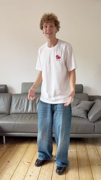 Balti marškinėliai su vyšnios piešiniu ant krūtinės, laisvi mėlyni džinsai, juodi batai. Stovi gyvenamajame kambaryje su pilku sofa ir medinėmis grindimis.