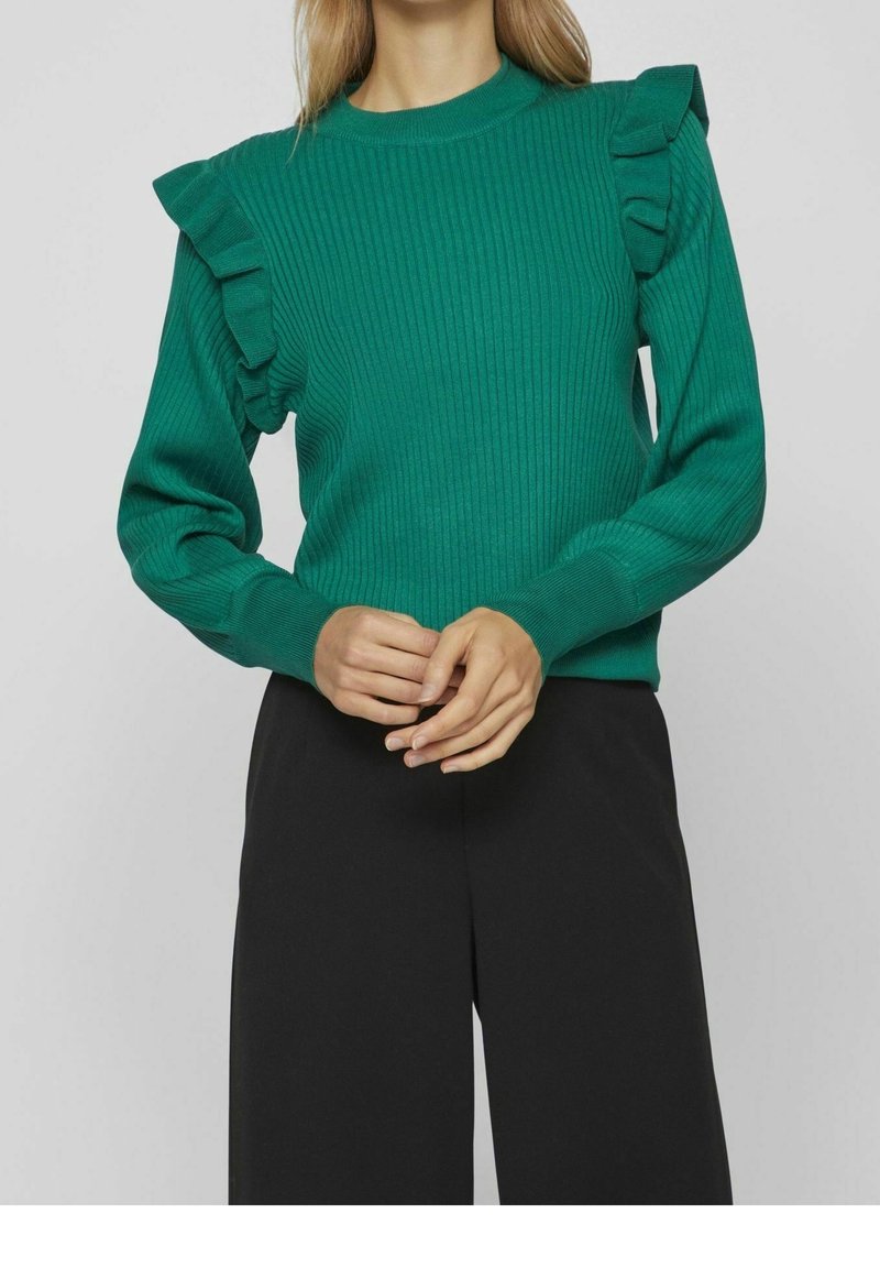 Grön ribbad stickad tröja med volanger på axlarna. Långa ärmar och rundad halsringning. Tillsammans med svarta byxor.