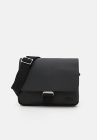 Svart messengerbag laget av teksturert materiale med klafflukking, justerbar stropp og metallbeslag. Har en logo detaljer på forsiden.