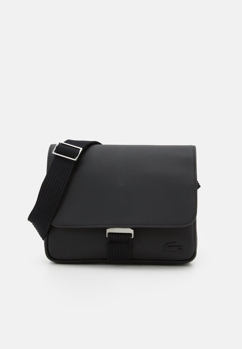 Svart messengerbag laget av teksturert materiale med klafflukking, justerbar stropp og metallbeslag. Har en logo detaljer på forsiden.