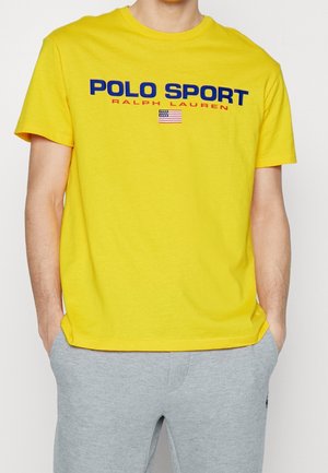 Żółta koszulka z krótkim rękawem z napisem „POLO SPORT RALPH LAUREN” i małą amerykańską flagą, noszona z jasnoszarymi spodniami, ręce w kieszeniach.