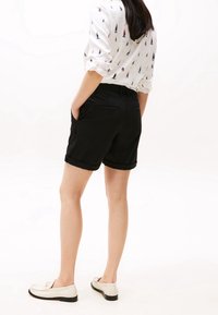 Sorte bomuldsshorts med opsmøgede afslutninger, to sidelommer og baglommer, båret med en mønstrede hvid skjorte og lyse loafers.