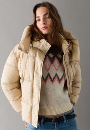 Junge Frau trägt eine beige Steppjacke über einem gemusterten Strickpullover und blauen Jeans, steht vor einem schlichten grauen Hintergrund.