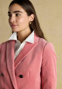 Naisel, kellel on pikad pruunid juuksed, on kuldsed rõngas kõrvarõngad, valge krae ja roosa kordoniga kahe nupuga bleiser, mis on asetatud beežile taustale.