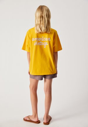 Kind mit blondem Haar, das ein gelbes T-Shirt mit dem Text "american vintage", graue Shorts und braune Flip-Flops trägt und mit dem Rücken zum Betrachter steht.
