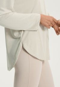 Világos bézs hosszú ujjú felső, ívelt aljjal és oldalslókkal, világos bézs leggings-szel párosítva. Puha anyag, sima textúrával.