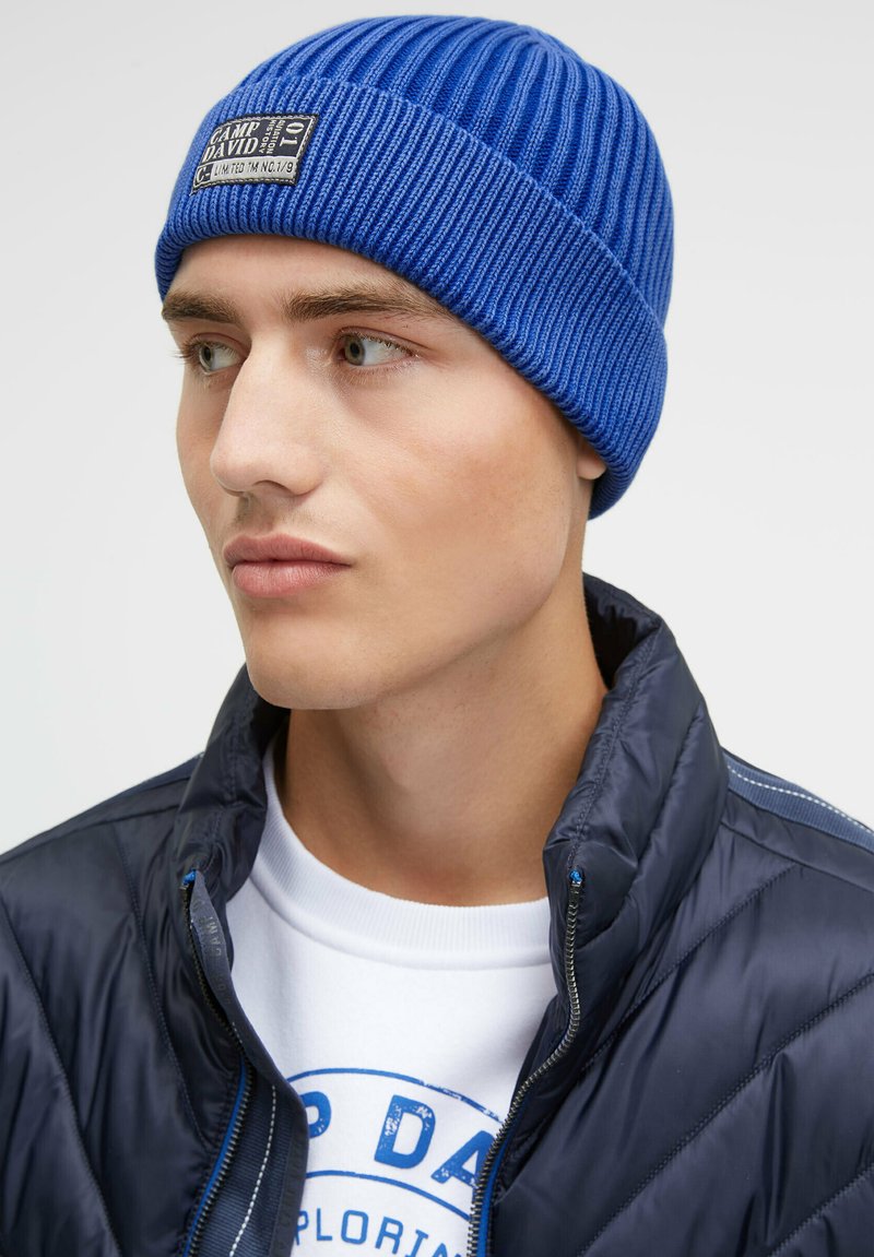 Blue ribbed knit beanie with a sewn label reading "CAMP DAVID." Worn with a navy puffer jacket and white sweatshirt beneath. Neutral background.