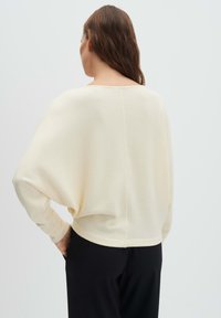 Frau mit langen braunen Haaren, die einen locker sitzenden, cremefarbenen Pullover und schwarze Hosen trägt, von hinten vor einem schlichten Hintergrund gezeigt.