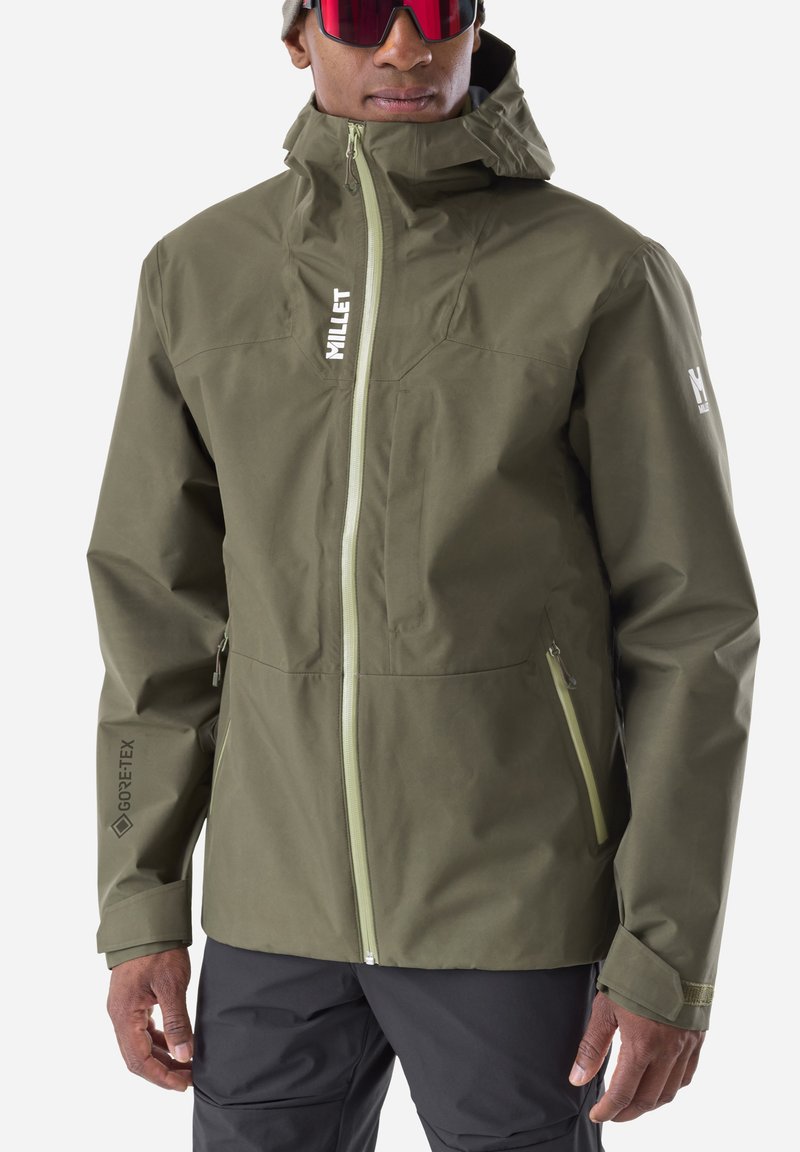 Man wearing olive green waterproof jacket with front zipper, zippered side pockets, and Gore-Tex logo on sleeve, along with dark pants and red sunglasses.