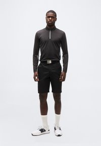 Νεαρός άνδρας που στέκεται, φορώντας μαύρο μακρυμάνικο μπλουζάκι με φερμουάρ, μαύρα σορτς με ζώνη, λευκές κάλτσες και λευκά αθλητικά παπούτσια, πάνω σε απλό φόντο.