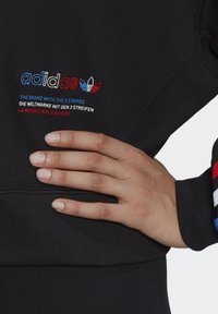 Svart sweatshirt med broderad "adidas"-logotyp i rött, blått och vitt. Har rund halsringning och färgglada randiga accenter på ärmarna.