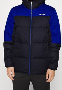 Puffer bunda v námořnickém a modrém barevném provedení, s zipovým zapínáním, vysokým límcem a logem na hrudi. Měkká, prošívaná textura.