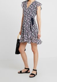 Blommig omlottklänning i marinblått med rosa och vita blommor. Lättviktigt tyg, volangärmar och knytmidja. Bärs med svarta sandaler och en väska.