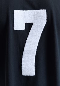 Écusson blanc texturé du numéro 7 sur un fond en tissu foncé, ressemblant à un maillot de sport ou un vêtement de style varsity.