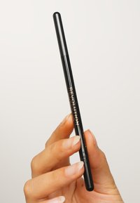 Matita eyeliner nera tenuta da una mano, caratterizzata da un design elegante con una finitura lucida e una punta arrotondata, etichettata "REVOLUTION" in oro.