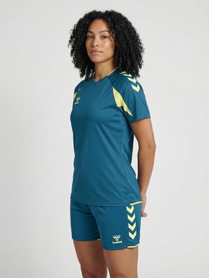 Kvinde med krøllet hår iført en sportsjersey og shorts i teal og gul, stående med armene bag ryggen mod en ensfarvet baggrund.