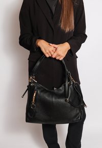 Sac à main en cuir noir avec une finition texturée, doté de quincaillerie dorée, d'une sangle détachable et d'un charme en forme d'ours. Le design est doux et décontracté.