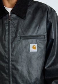 Giacca nera sintetica con colletto in velluto a coste, chiusura frontale con zip e tasca a patta con logo Carhartt in arancione e bianco.