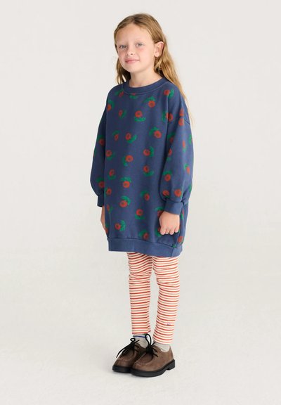 Jeune fille aux longs cheveux blonds portant un sweat-shirt bleu à fleurs oversize, des leggings rouges et blancs rayés, et des chaussures marron à lacets.