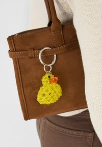 Gul gummiand nøgleholder med orange næb, fastgjort til en brun ruskinds håndtaske. Håndtasken har en solid sølvhardware-ring.