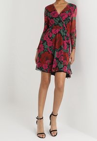 Blumenkleid mit langen durchsichtigen Ärmeln, das große rote und pinke Blumen auf einem dunklen Hintergrund zeigt, sowie einen weit ausgestellten Saum. Schwarze Riemchen-Highheels.