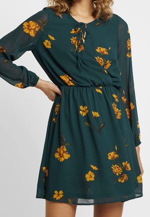 Robe fleurie en chiffon vert foncé, ornée de fleurs jaunes, avec un décolleté en forme de goutte d'eau à nouer, une taille élastique et des manches longues.