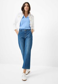 Lichtblauw V-hals top, witte denimjas met gouden knopen, hooggetailleerde blauwe jeans en witte sneakers met bruine zolen.