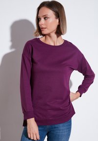 Maglietta lunga viola a maniche lunghe con scollo rotondo e vestibilità rilassata. Realizzata in tessuto morbido, presenta polsini a costine e spacchi laterali all'orlo.