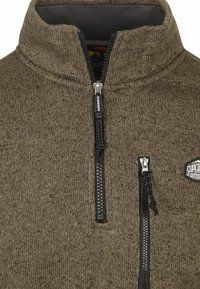Bruine fleecetop met hoge kraag, halve ritssluiting aan de voorkant en contrasterende zwarte ritsen. Heeft een logopatch op de borst.