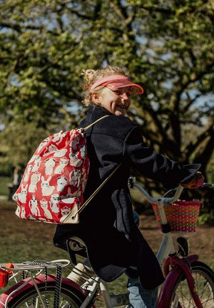 Rode trekkoordrugzak met patronen van een konijn en een kat, gemaakt van stof. Fietsser met een lichtblauwe fiets en een roze mand; zichtbare begroeiing op de achtergrond.