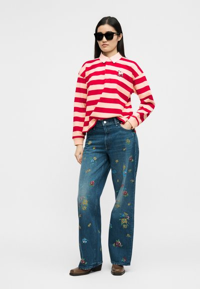 Γυναίκα που φοράει μαύρα γυαλιά ηλίου, ροζ και κόκκινο ριγέ μακρυμάνικο πόλο, μπλε τζιν με λουλουδάτη κεντητική και καφέ παπούτσια, στέκεται με το χέρι στην τσέπη.