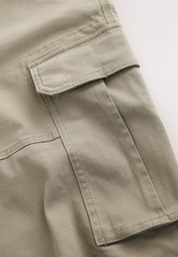 Un tissu kaki à la texture lisse présente une grande poche plaquée avec des coutures détaillées. Le design comprend un rabat plié sur la poche.