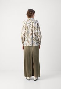 Camicia a maniche lunghe con fantasia floreale in tessuto leggero, abbinata a una lunga gonna verde oliva con spacco posteriore e scarpe bianche basse.