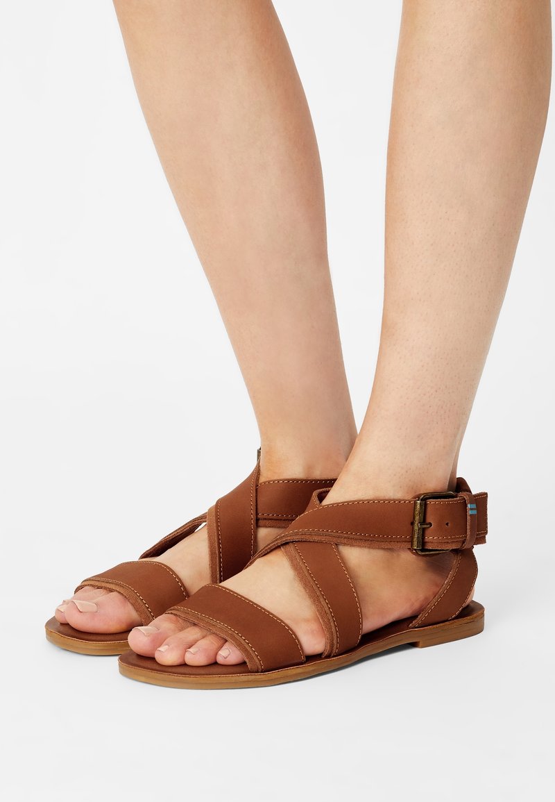 Pieds portant des sandales plates en cuir marron avec des lanières croisées et une fermeture à boucle à la cheville sur un fond blanc.
