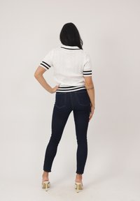 Frau steht mit dem Rücken zur Kamera, trägt ein weißes Strickoberteil mit kurzen Ärmeln und schwarzen Streifen sowie dunkle enge Jeans und hochhackige Schuhe.