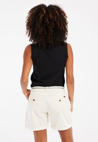 Pantalones cortos de color claro con dobladillo enrollado, cinturón blanco con rayas verdes y dos bolsillos traseros con botones; combinados con una blusa sin mangas negra.