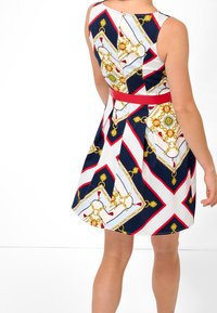 Hihaton, vartalonmyötäinen mekko, jossa rohkea, monivärinen geometrinen kuviointi; päävärit ovat laivastonsininen, valkoinen ja punainen, ja mekkoa korostavat kultaiset yksityiskohdat.